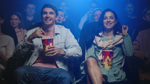 Cheerful friends laughing in cinema. Happy couple having fun in movie theater. Beautiful woman and handsome man watching movie together indoor.