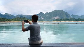 Back View of Man Filming Blog or Vlog, Video Chat on Mobile Phone at Amazing Lake with Green Mountains. Travel Guy Videographer in Nature Scenic. Cheow Lan Lake in Khao Sok Park in Thailand.