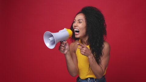 Surprised laughing african american young woman in basic yellow tank top posing isolated on red background studio. People lifestyle concept. Screaming in megaphone say wow pointing index finger aside