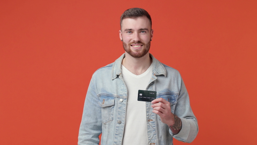 Excited funny bearded young man 20s in white t-shirt denim jacket isolated on orange background studio. People lifestyle concept. Point index finger on credit bank card showing thumbs up like gesture.