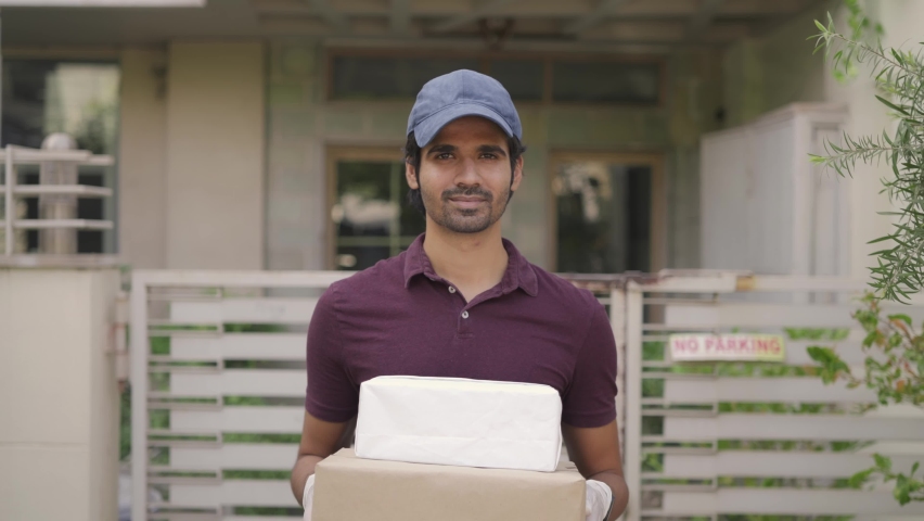 Track out shot of a young attractive Indian Asian delivery man executive holding a parcel in hand, standing with a smile on face in front of a house looking at the camera Royalty-Free Stock Footage #1066656997