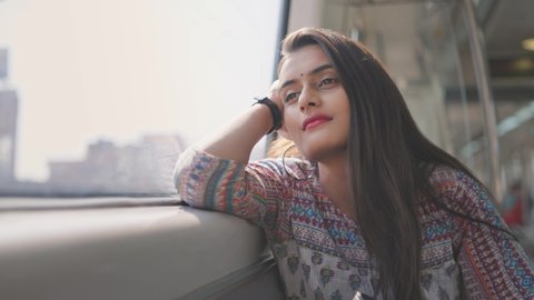 A close shot of a beautiful Indian young woman solo traveler in traditional salwar kurta sitting in a moving metro train leaning on a window and looking outside 