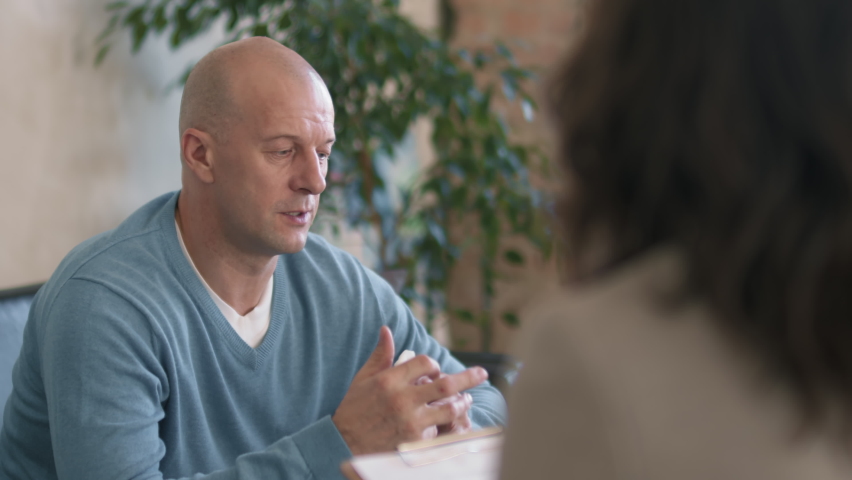 Tracking over the shoulder shot of unrecognizable female therapist writing notes on clipboard and listening to emotional middle-aged man talking about his problems during session