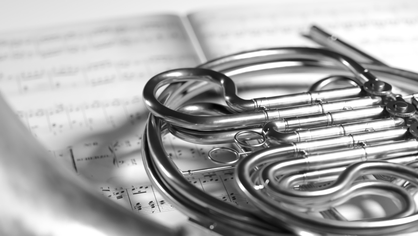 Black and white close up shot of a french horn resting on music note sheets. Slow camera movement. A brass instrument made of shiny tubing used by players in proffesional orchestra or band.