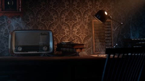 Old room with vintage wooden desk and chair. Dark interior with warm light. Cobwebs coated old-fashioned radio, books and abacus. Paper and pen on a table lightened by a lamp. Atmosphere of mystery.
