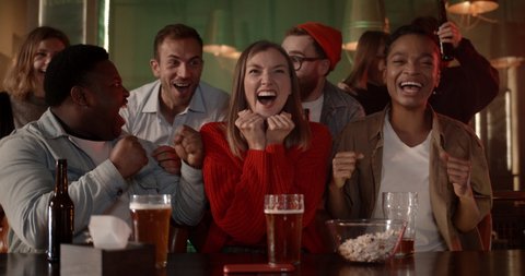 Joyful diverse people celebrating and smiling while watching sports game. Happy young friends rejoicing and shouting while supporting their favourite team and having beer in pub.