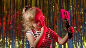 Trendy stylish child kid blogger smiling cheerfully, recording video blog vlog on smartphone with tripod. Fashion girl in futuristic night club with red neon lights making selfie call on mobile phone