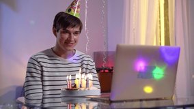 Happy millennial birthday man waving and talking in video chat on laptop showing thumbs up. Candles burning on cake. Portrait of positive Caucasian guy celebrating party remotely online.