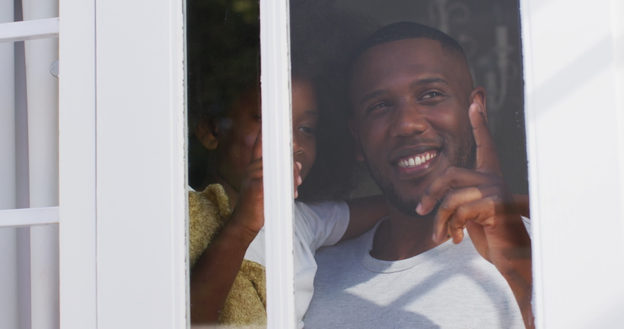 African american daughter holding teddy bear looking through window with her father. having fun staying at home in self isolation during quarantine lockdown.