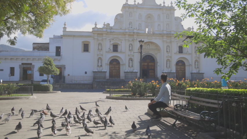 Hispanic young man sitting in the central park of Antigua Guatemala while pigeons fly in the middle of the plaza -young man in colonial city looking towards the cathedral  | Shutterstock HD Video #1066671901