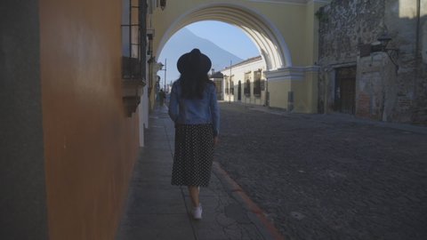 Young Hispanic woman walking in emblematic street of Antigua Guatemala below Arch of Santa Catalina - young man walking through colonial city early in the morning 