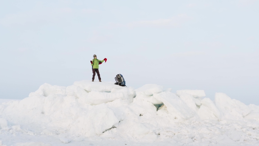 Aerial panoramic view of a happy woman dancing and waving the red scarf on big icy blocks against the blue sky. Seaside. Frozen sea is on the background. Winter