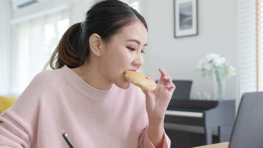 Young attractive beautiful asian female hungry eat doughnut take away snack food with full mouth look at computer notebook at home in busy work from home multitask unhealthy meal lifestyle concept. | Shutterstock HD Video #1066672666