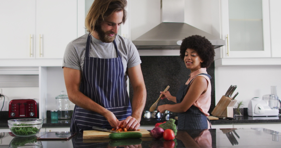 Mixed race couple wearing aprons cooking food together in the kitchen. staying at home in self isolation in quarantine lockdown | Shutterstock HD Video #1066673533