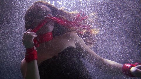 Portrait of an attractive woman in a red mask, with a whip in her hands, a collar around her neck, she is under water creating a vortex of bubbles.