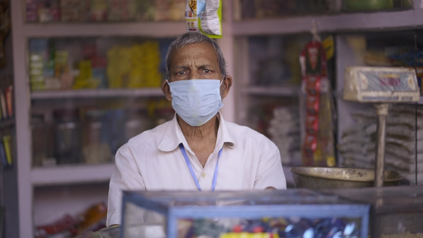 Frontal shot of an Indian Asian adult grocery shopkeeper or owner wearing a protective mask on a face sitting on a shop counter looking at camera during the hard times of Covid 19 epidemic Royalty-Free Stock Footage #1066681519