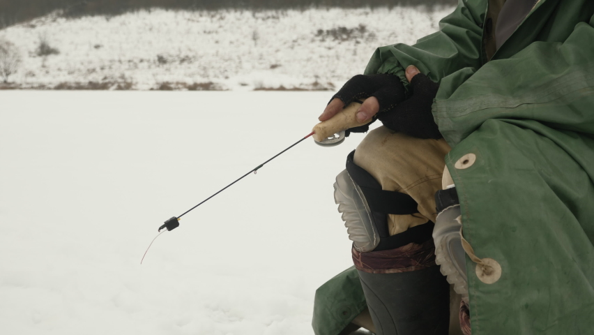 Man in winter extreme clothes with fishing rod catches fish in ice hole. Fisherman at winter fishing on frozen lake or river