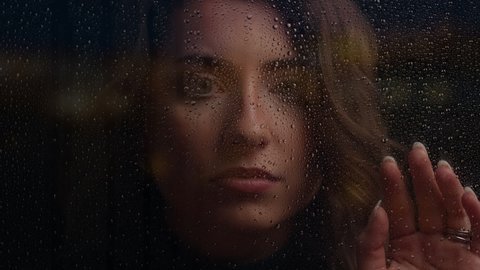 Close up of woman looking at camera with hand on window on rainy night. Drops of water on window are in focus. Cinematic