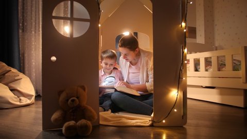 Dolly shot of young mother reading book to her little son while sitting in small toy house. Concept of child education and family having time together at night.