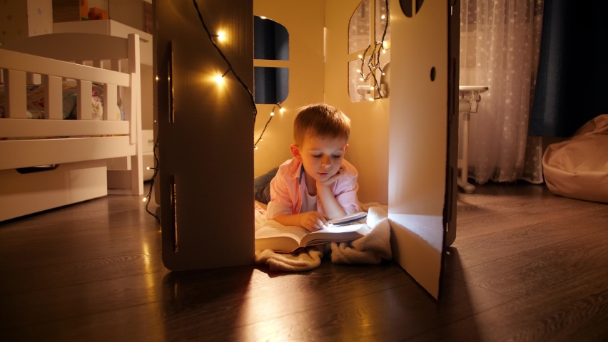 Happy smiling boy lying on floor at hos toy house or tent and reading with flashlight at night. Concept of child education and reading in dark room. | Shutterstock HD Video #1066699480