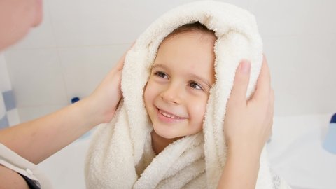 Portrait of cute smiling boy with wet hair wiping and drying with soft towel after having bath and looking on caring mother. Concept of child hygiene and health care at home