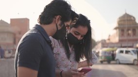 A young Indian male and female friends with face protective mask looking into a mobile phone, smiling and talking to each other standing by a city pedestrian walkway with busy road traffic around 