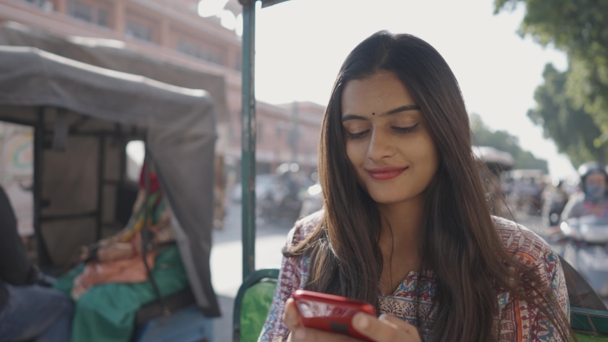 A close shot of a young beautiful Indian female sitting and using a mobile phone to type a text message while sitting in a moving local electric auto-rickshaw going through a busy city street Royalty-Free Stock Footage #1066700293