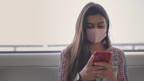 Close shot of a young attractive Indian female sitting on a moving city metro train with a protective mask on her face and typing a text message on a mobile phone during Covid 19 epidemic or pandemic 