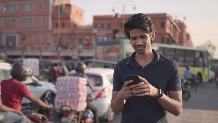 close view shot of young attractive Indian male using a mobile phone to type a text message with smile on face walking on a footpath alongside busy road traffic and market place Royalty-Free Stock Footage #1066700404