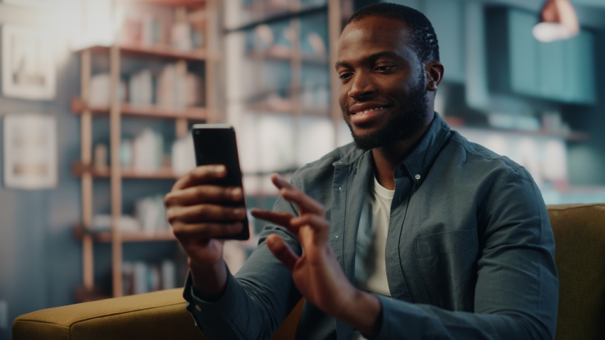Excited Black African American Man Using Smartphone while Resting on a Sofa in Living Room. Happy Man Smiling at Home and Chatting to Colleagues and Clients Over the Internet. Using Social Networks.