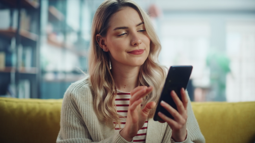 Beautiful Caucasian Female Using Smartphone in Stylish Living Room while Resting on a Cozy Couch Sofa. Young Woman at Home, Browsing Internet, Using Social Networks, Having Fun in Flat.
