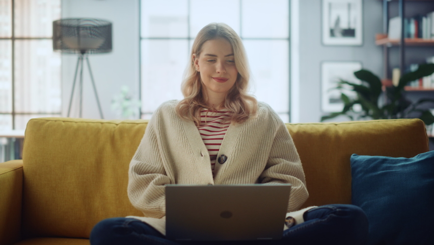 Beautiful Caucasian Specialist Working on Laptop Computer at Stylish Home Living Room while Sitting on a Cozy Couch Sofa. Freelance Female Browsing Internet, Using Social Networks, Having Fun in Flat.