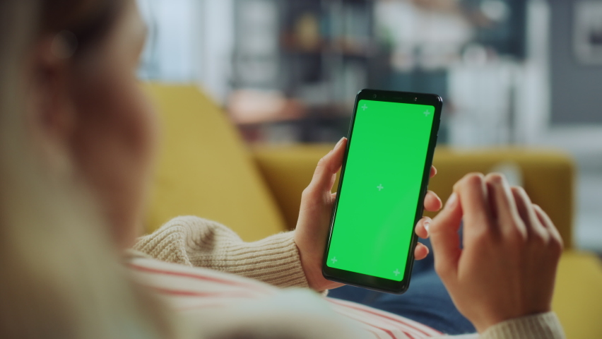 Beautiful Authentic Female Using a Smartphone with Green Screen Mock Up Display at Home Living Room while Lying on a Couch Sofa. She's Browsing the Internet and Checking Videos on Social Networks.