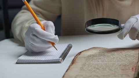 Researcher in cotton gloves examines antique book with magnifying glass. Scientific translation of ancient literature. Studying manuscript with ancient writings