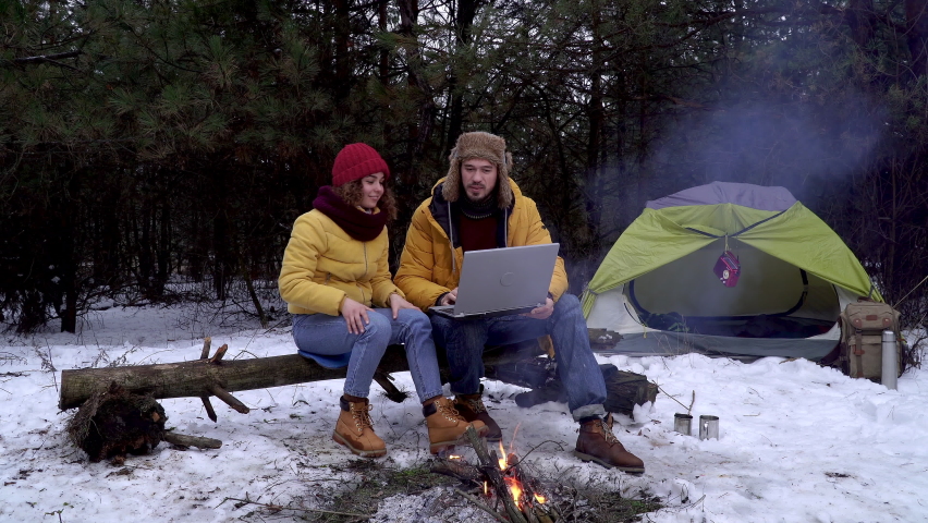 Young man and woman with a laptop near a campfire in the winter forest. They communicate with friends using the Internet.