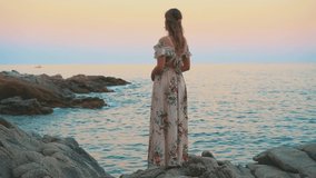 Silhouette of woman tourist stands on shore stone coast adriatic blue sea. Beautiful sunset pink yellow sky. Happy girl dream, enjoy nature, view water Costa Brava beach Lloret de Mar. Back long dress