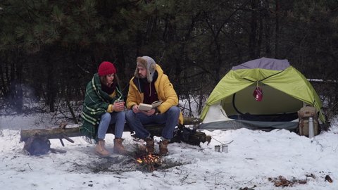 Young man and woman read a book by the fire in the winter forest. Tourists travel together. Travel and adventure concept.