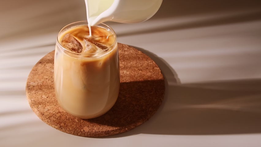 Milk cream is pouring into a iced coffee. Cold coffee drink glass with ice and milk. | Shutterstock HD Video #1066723705