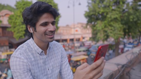A young Indian male office worker sitting on a rooftop of a building and having tea while having a conversation over a video call on a mobile phone with busy market road traffic of evening rush hour 