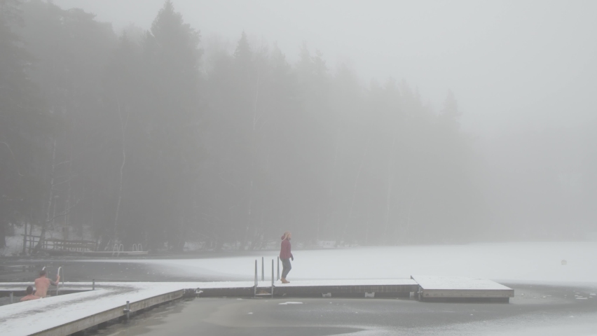 Tracking sideways shot of a Scandinavian woman walking along boardwalk over lake in wintery and moody national park in Finland with steam rising from the water.