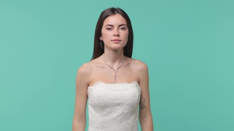 Confused puzzled bride young woman 20s in beautiful white wedding dress posing isolated on blue turquoise color background studio. Wedding concept. Put hand prop up on chin spreading arms say oops no