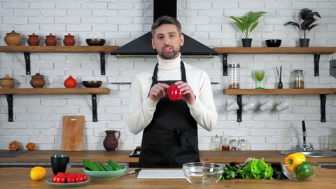 Man chef food blogger in black apron looking camera tells teaches records remote online video culinary webinar master class course in home kitchen, vegetables ingredients cooking salad on table