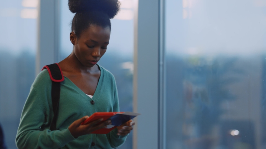 Young african casual lady using tablet computer reading e-book application walking near window in business center. Airport terminal building. Departure waiting room.