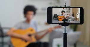 Focus on smartphone, close-up image on mobile phone afro american guy man teacher recording video lesson for blog for students teaches to play guitar musical instrument remotely, broadcasting concept