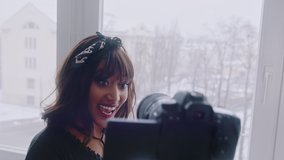 Cheerful young African American female blogger holding camera and recording video for vlog while standing near the window. High quality 4k footage