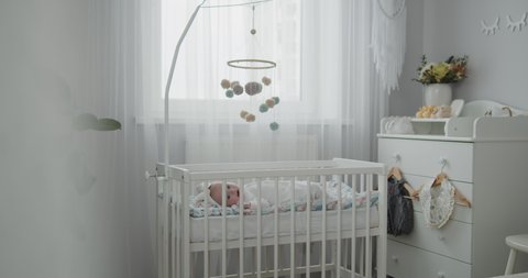 Stylish bright modern baby room interior. Cute newborn toddler lying in the cradle. Crib mobile hanging above, booties on a changing table. Concept of childhood, new life, parenthood.
