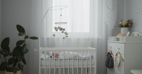 Stylish bright modern baby room interior. Cute newborn toddler lying in the cradle. Crib mobile hanging above, booties on a changing table. Concept of childhood, new life, parenthood.