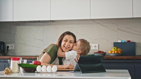 slow motion: little child hangs around mother's neck, daughter with long hair cheerfully hugs with both hands, stand in kitchen, cook lunch with vegetables and fruits, guided by recipes from Internet