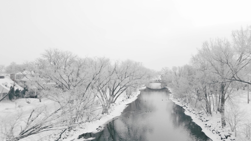 Aerial video following a river lined on both sides by white snow and frost covered trees revealing a foot bridge and a frozen barren lake.