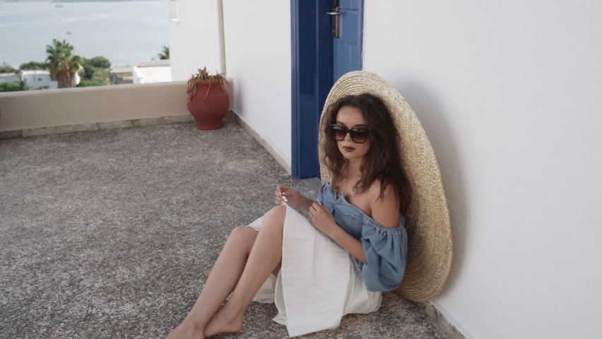 Middle shot of young woman sitting down on the ground in a light white dress and watching a beautiful sea scenery.